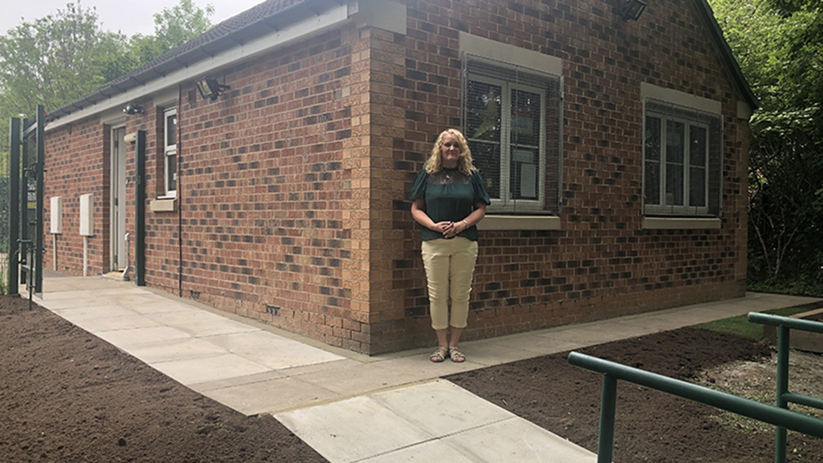 Woman standing outside a 1 storey brick building, with grills on the windows.